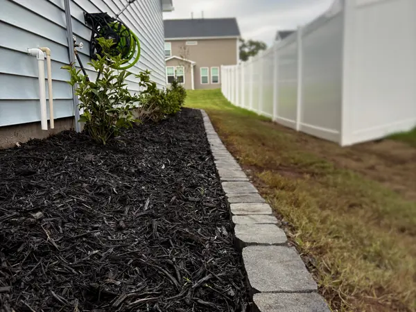A long mulch bed with a stone paver border running beside a white fence.
